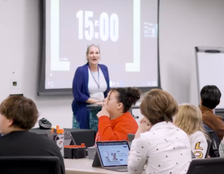 Students seated around tables with tablets listen to a presenter at the front of a classroom during a Digital Citizenship Symposium session.