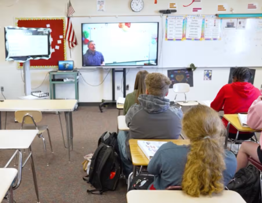A classroom of students sit at desks facing the front of the room while a distance learning teacher stands in frame in an interactive display screen presenting a lesson 