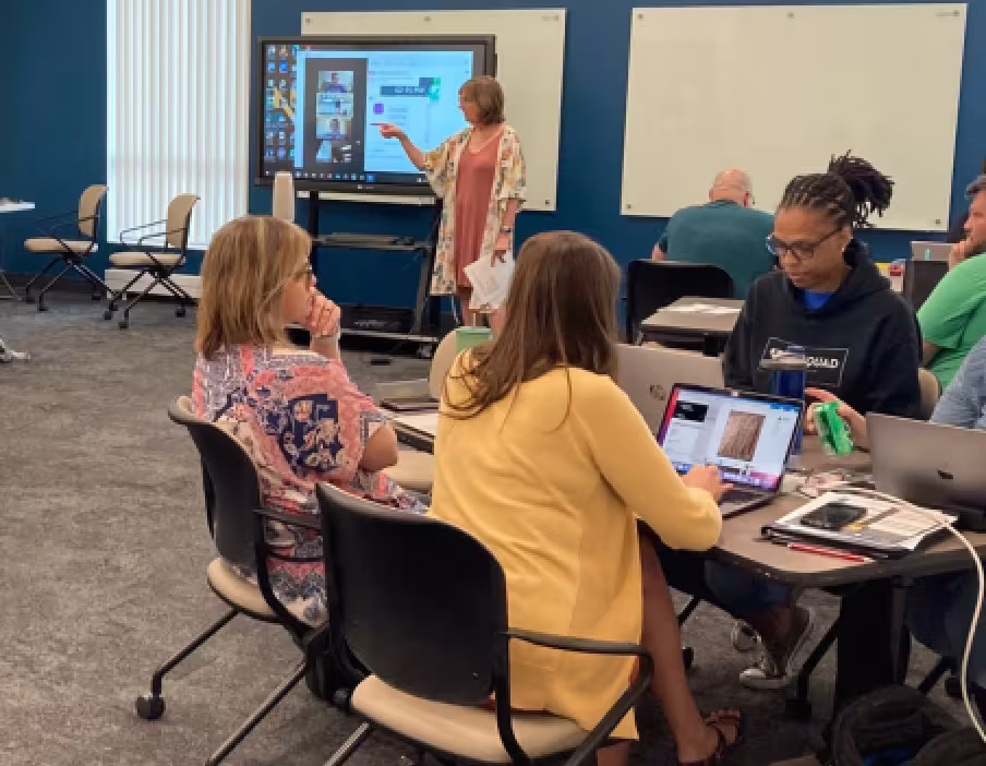 An ESU educator stands at the front of a classroom or teachers pointing to content on an interactive display while adult learners sit at tables working on laptops and discussing in small groups.