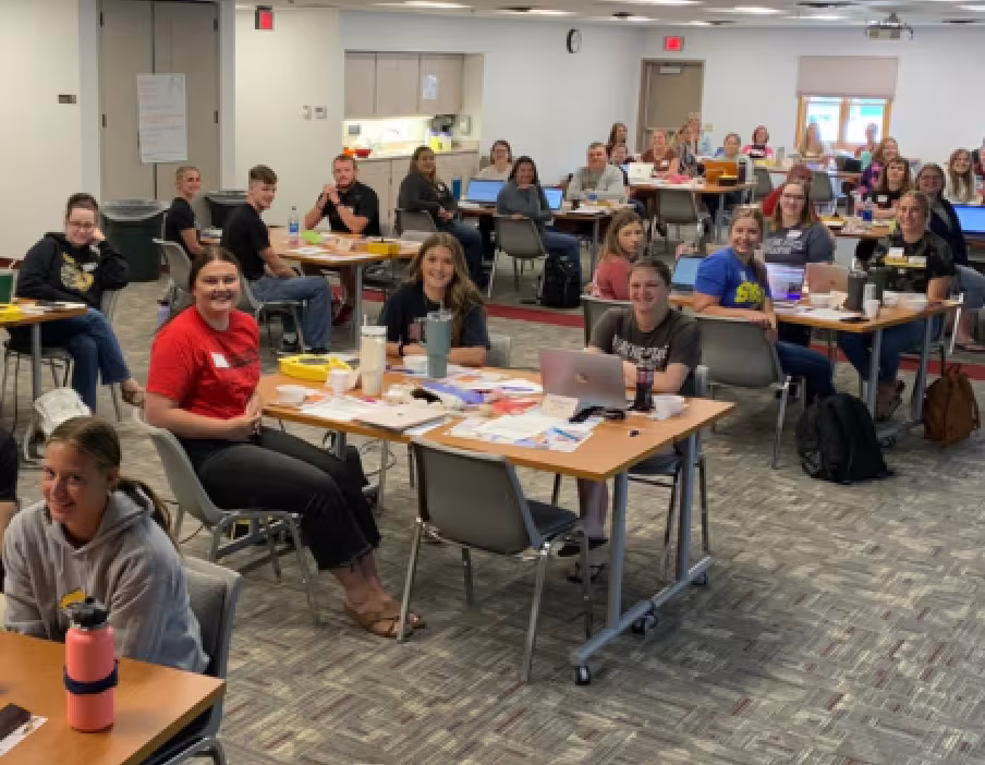 Group of adult educators seated at tables in a training room, smiling and looking toward the camera, with laptops, notebooks, and papers on the tables.