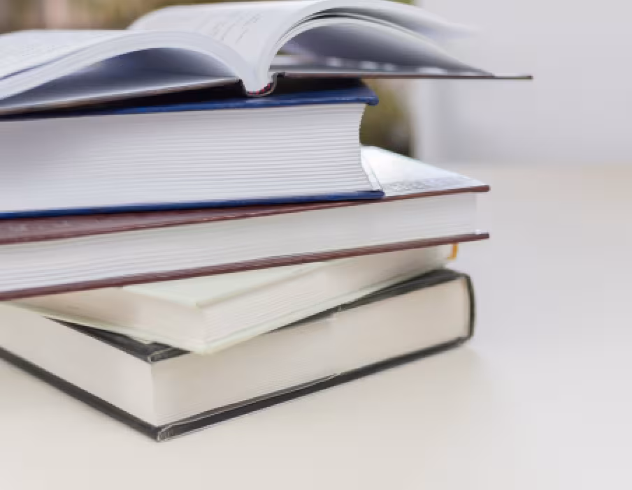Stack of hardcover books on a table with one book open on top, pages slightly curved.