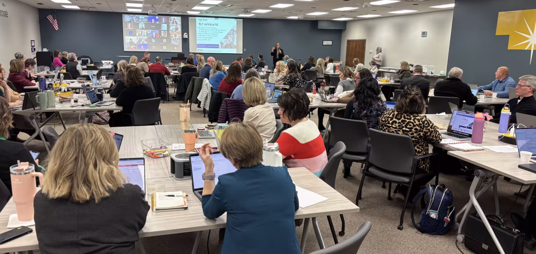 Large group of educators are seated at tables during a session at the ESUPO statewide convening. Those educators are using laptops and also listening to a presenter at the front of the room near two projected presentation screens.