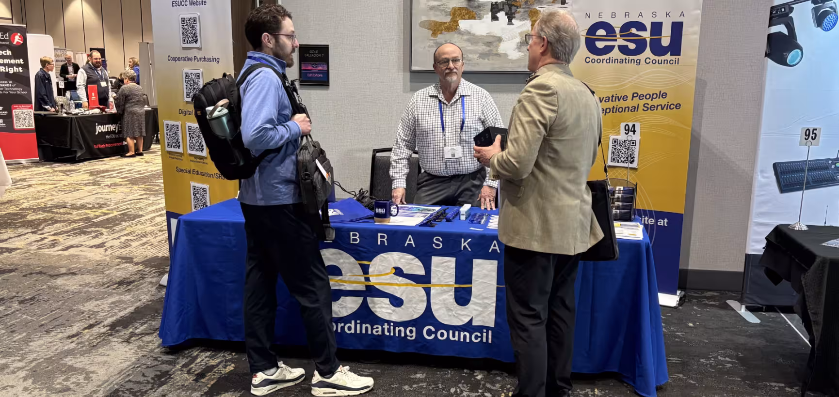 Three men talk at a conference exhibit booth for the Nebraska ESU Coordinating Council, with banners, a table display, and QR codes visible behind them.