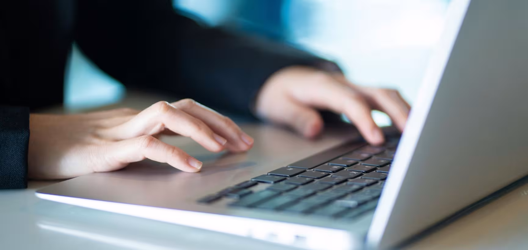 Close-up of a person’s hands typing on a laptop keyboard, with fingers positioned over the keys and the screen out of focus in the background.